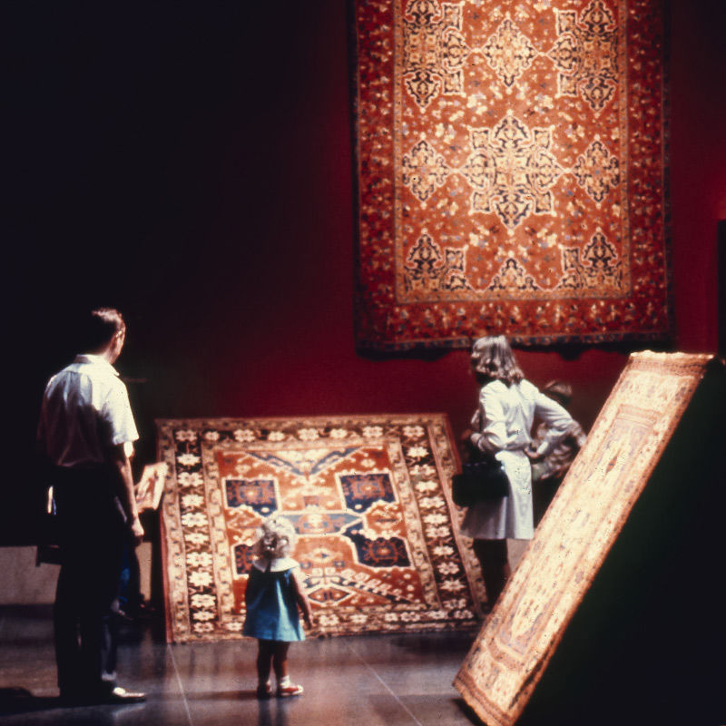 A man, small girl, woman and young boy stand around facing a large Persian-style rug in dark reds and browns displayed on a slanted ramp on the floor. The room is painted a dark maroon color with bright spotlights over each displayed carpet. Another similar large red rug with vegetal arabesque patterns and motifs hangs on the wall to the right of the group.